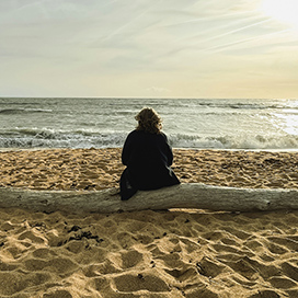 A woman sitting on a piece of driftwood on a beach, looking out to sea